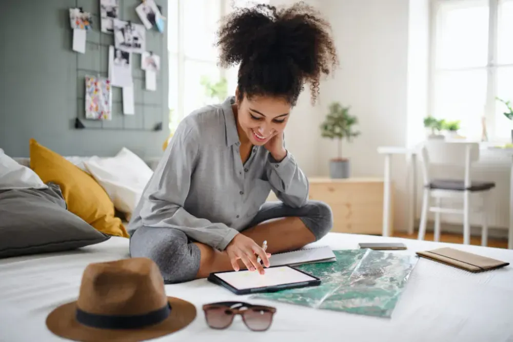 Woman sitting on a bed and working on tablet surrounded by a map, sunglasses, a hat, and small pocket books